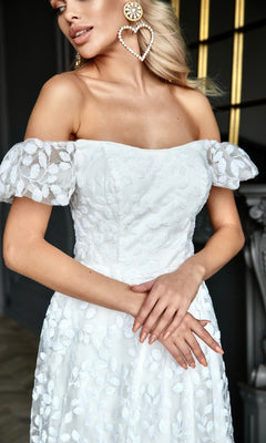 A woman stands indoors with arms crossed, wearing the Long White Lace Formal Dress: Tina Holly TK023W, featuring an off-the-shoulder design and sheer sleeves, accessorized with large heart-shaped pearl earrings.