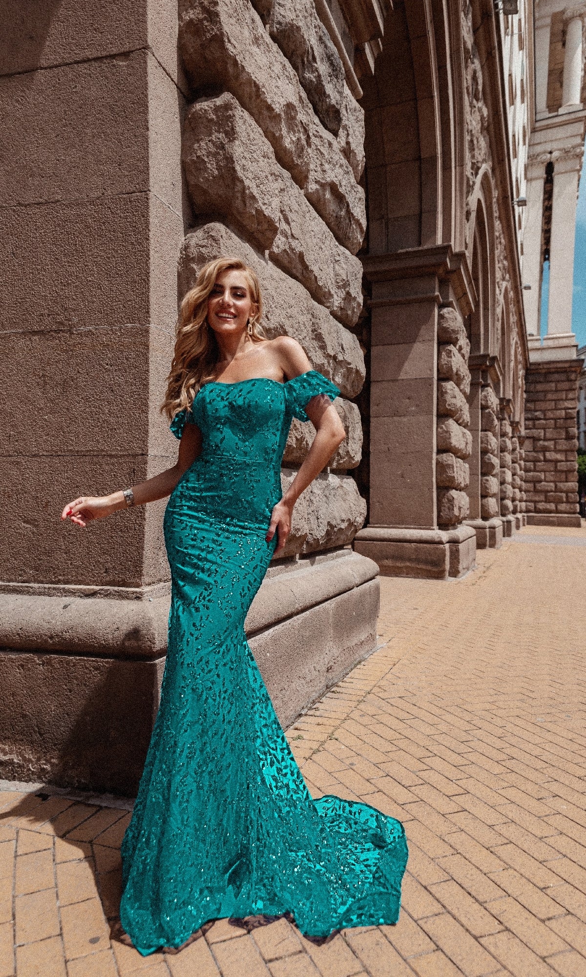 A woman in the Long Glitter-Lace Prom Dress, Tina Holly TW024, stands smiling against a stone building with arched architecture on a sunny day.