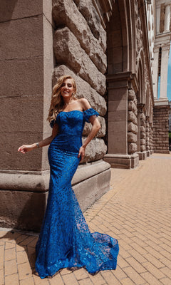 A woman wearing the Long Glitter-Lace Prom Dress (Tina Holly TW024), a fitted off-the-shoulder gown, smiles beside a stone building with arched columns on a sunny day.