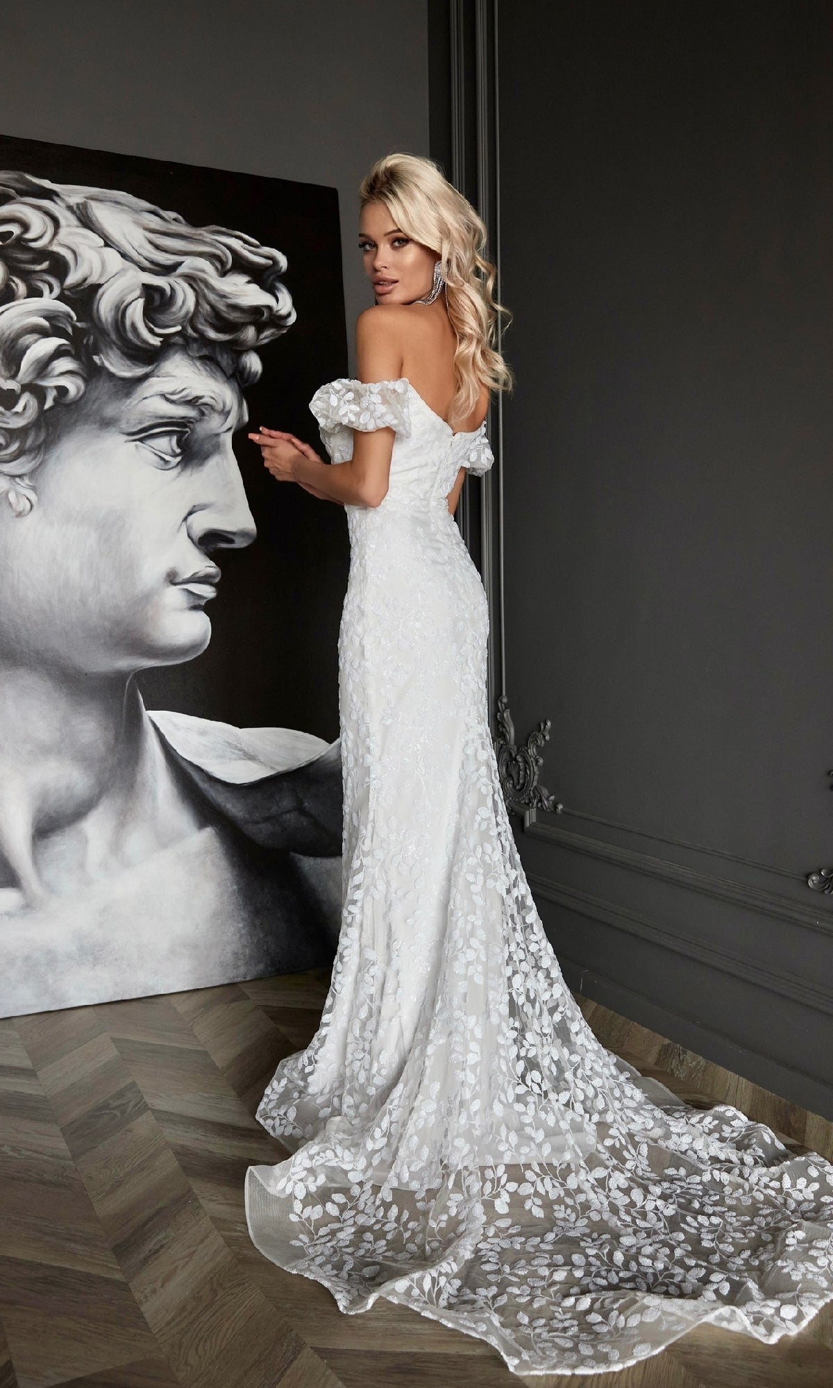 A woman in the Off-Shoulder White Formal Dress: Tina Holly Tk024W with a floral pattern stands near a large classical sculpture artwork in a dimly lit room with wooden floors.
