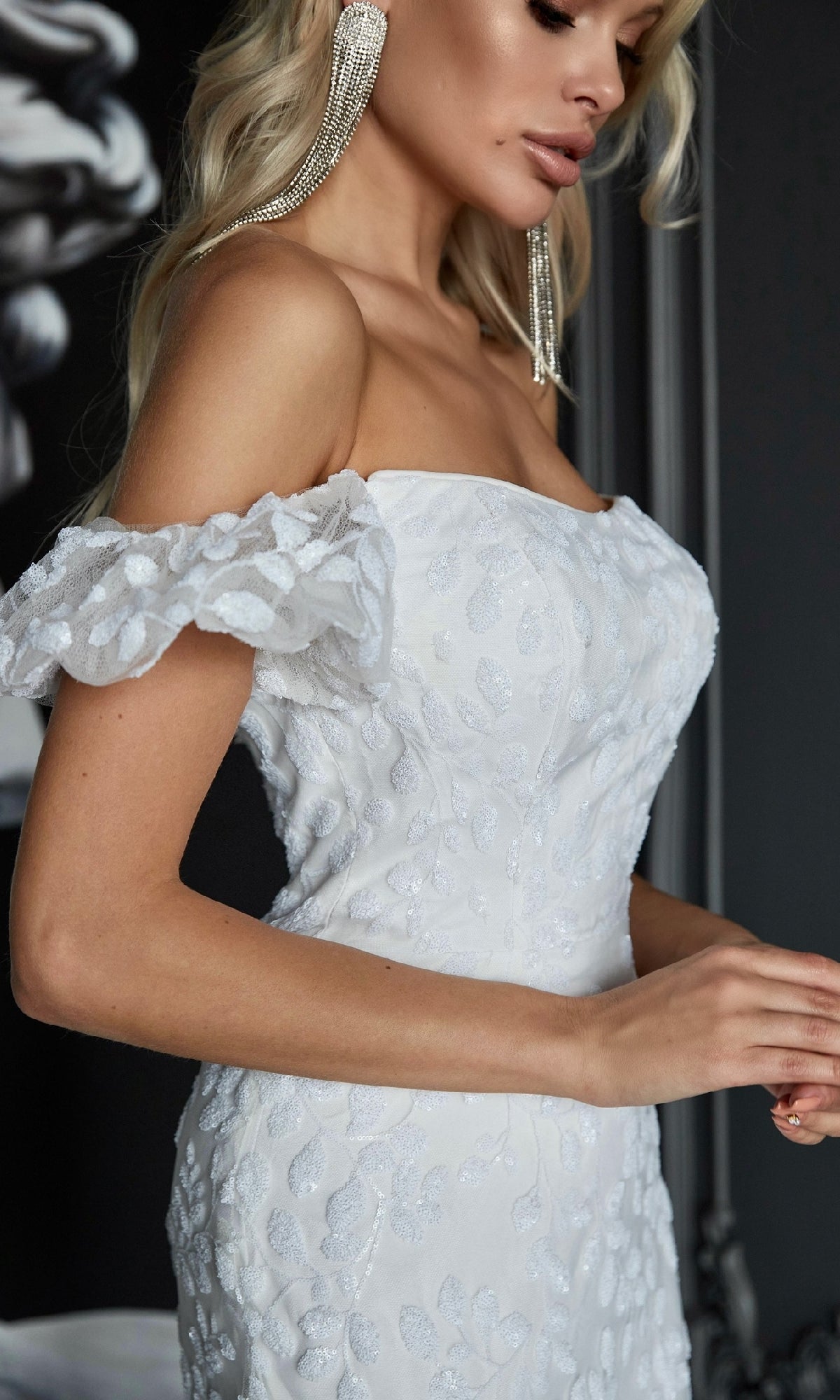 A woman stands indoors against a dark background, wearing the Off-Shoulder White Formal Dress: Tina Holly TK024W and large earrings.