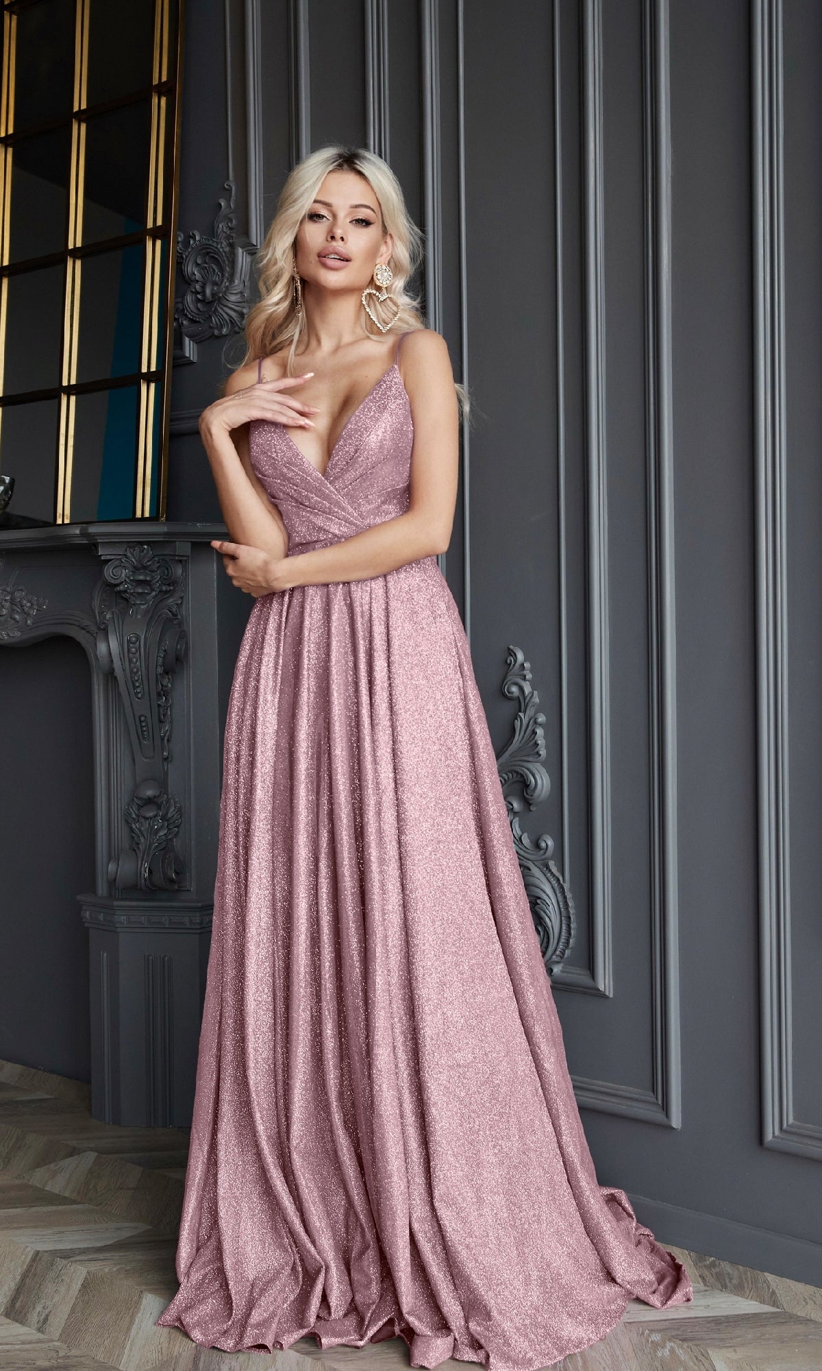 A woman poses elegantly with one hand on her waist in front of an ornate gray wall and fireplace, wearing the Long Glitter A-Line Prom Dress by Tina Holly TW079, featuring a shimmering finish and deep v-neck.