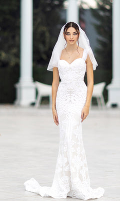 A woman in the Off-Shoulder White Bridal Gown: Tina Holly TW103, featuring lace and a veil, stands outdoors on a paved surface, looking at the camera.