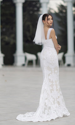 A woman in the Off-Shoulder White Bridal Gown: Tina Holly TW103 stands outdoors, looking over her shoulder, with white columns and blurred greenery in the background.