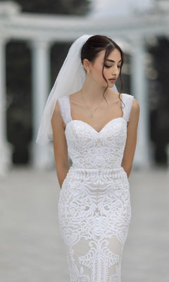 A bride wearing the Off-Shoulder White Bridal Gown: Tina Holly TW103 with lace and veil stands outdoors, looking down with her hands behind her back. Blurred marble columns are visible in the background.