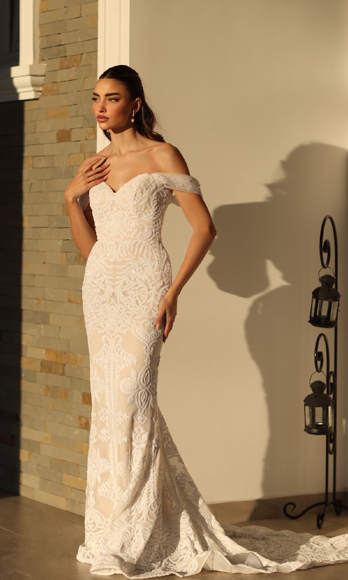A woman stands indoors near a stone wall and lanterns, wearing the Off-Shoulder White Bridal Gown: Tina Holly TW103, a fitted lace mermaid wedding dress.