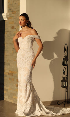 A woman stands indoors near a stone wall and lanterns, wearing the Off-Shoulder White Bridal Gown: Tina Holly TW103, a fitted lace mermaid wedding dress.