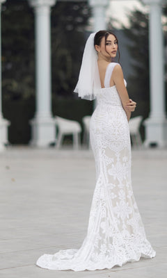 A woman wears the Off-Shoulder White Bridal Gown: Tina Holly TW103 with delicate lace and veil, standing outdoors on a paved area framed by elegant white columns in the background.