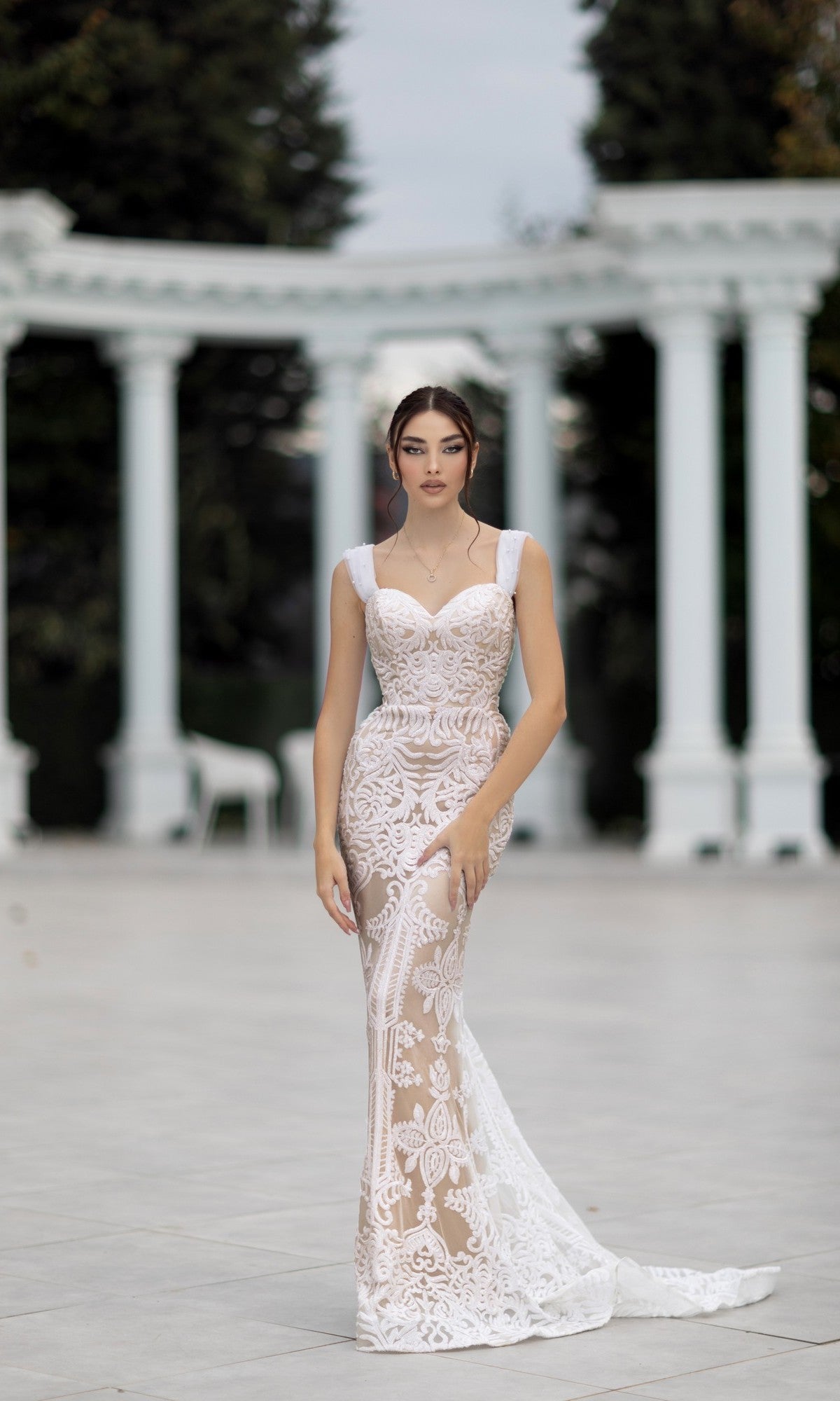 A woman stands outdoors in the Off-Shoulder White Bridal Gown: Tina Holly TW103, a fitted lace mermaid dress with sheer design. Elegant white columns and lush greenery provide a stunning backdrop.