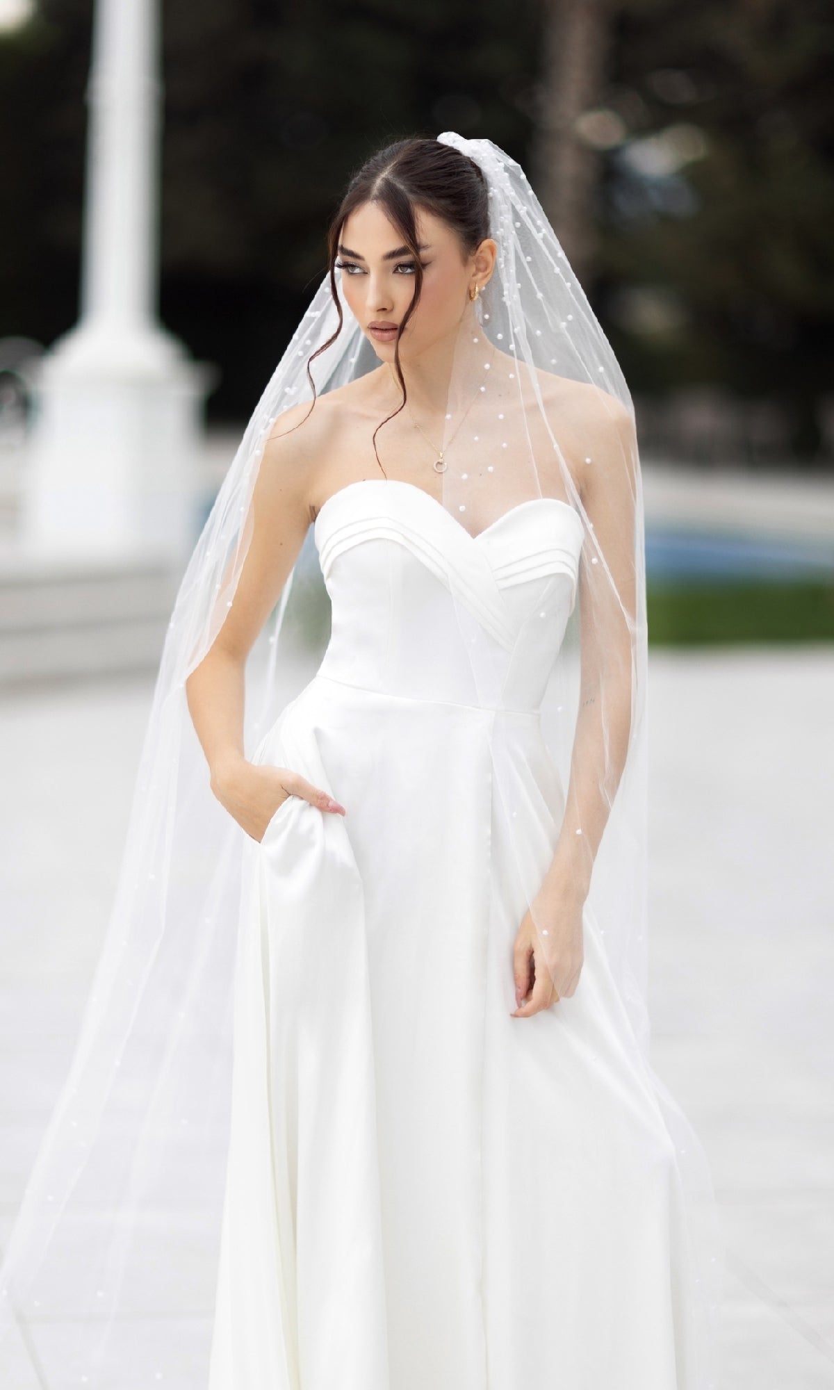 A woman stands outdoors in the Off-White Strapless Bridal Gown: Tina Holly TW105, featuring a sweetheart neckline, long pearl-accented veil, and one hand tucked in her pocket.
