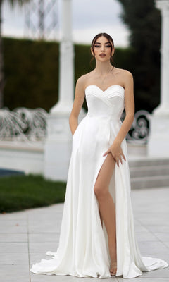 A woman wearing the Off-White Strapless Bridal Gown: Tina Holly TW105, featuring a thigh-high slit and sweetheart neckline, stands outdoors on a tiled surface, posing with one hand holding the dress.