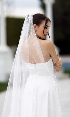 A woman in the Off-White Strapless Bridal Gown: Tina Holly TW105 and a sheer veil stands outdoors, gazing gracefully over her shoulder.