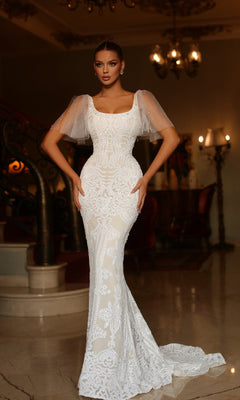 A woman stands indoors wearing the Tina Holly TW111 short sleeve white formal gown, featuring a fitted lace design with sheer flutter sleeves, in an elegant, dimly lit room with a curved staircase and ornate decor.