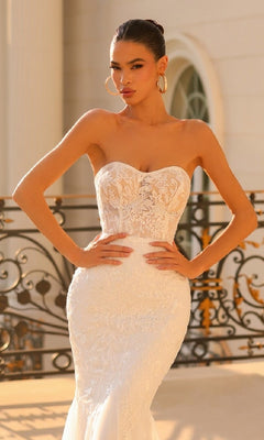 A woman stands outdoors with hands on hips, wearing the Off-White Strapless Bridal Gown: Tina Holly TW122, featuring a long silhouette and an embroidered-lace bodice, in front of an ornate railing.