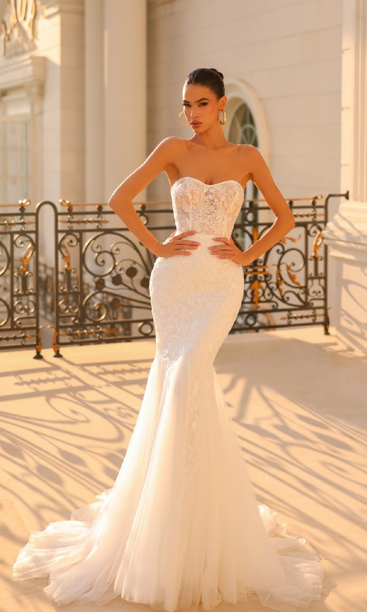 On a sunlit terrace with ornate railings, a woman wears the Off-White Strapless Bridal Gown: Tina Holly TW122, featuring an embroidered-lace bodice and elegant mermaid silhouette.
