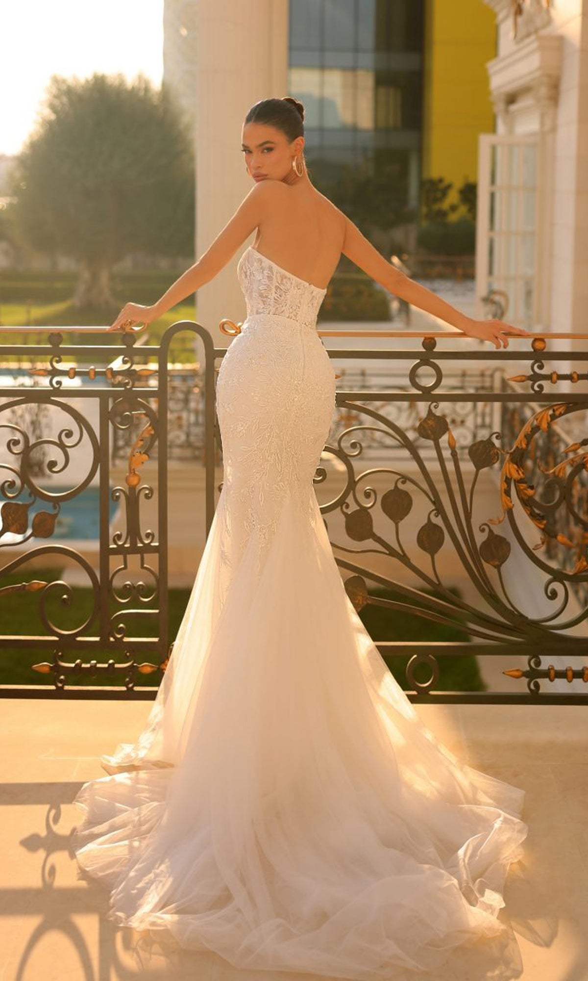 A woman in the Off-White Strapless Bridal Gown: Tina Holly TW122, featuring a fitted embroidered-lace bodice and long train, stands on a balcony and looks over her shoulder toward the camera.