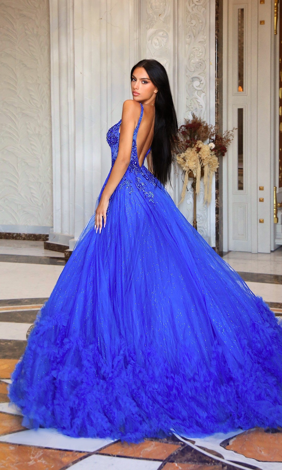 A woman stands indoors on a marble floor in an ornate room with white walls and dried florals, wearing the Tina Holly TW610 long prom dress featuring a bright blue hue, low back, and sheer corset bodice.