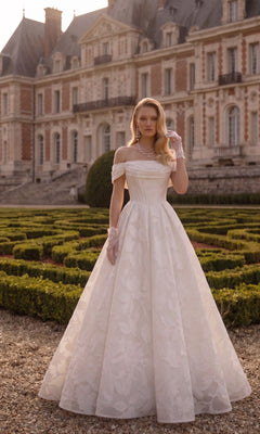 A woman in a Tina Holly TW623 long wedding dress, featuring an off-the-shoulder, floral-patterned organza jacquard gown and lace gloves, stands in front of a historic manor.