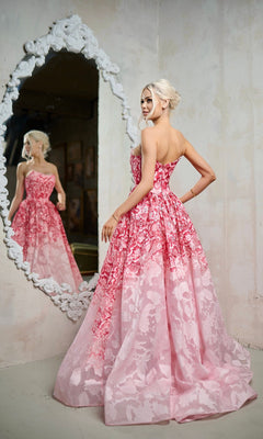 A blonde woman in the Tina Holly TW624 long formal dress stands before an ornate mirror, gazing at her reflection. The strapless A-line silhouette and soft pink floral organza jacquard fabric enhance her elegant pose.