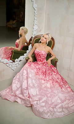 A woman in the Tina Holly TW624 long formal dress in floral pink sits on a green chair, her reflection visible in a large ornate mirror beside her.