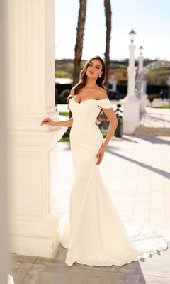 A woman in a Tina Holly TW628 wedding dress with an off-the-shoulder, sweetheart neckline, fitted bodice, and small train standing beside a white pillar.