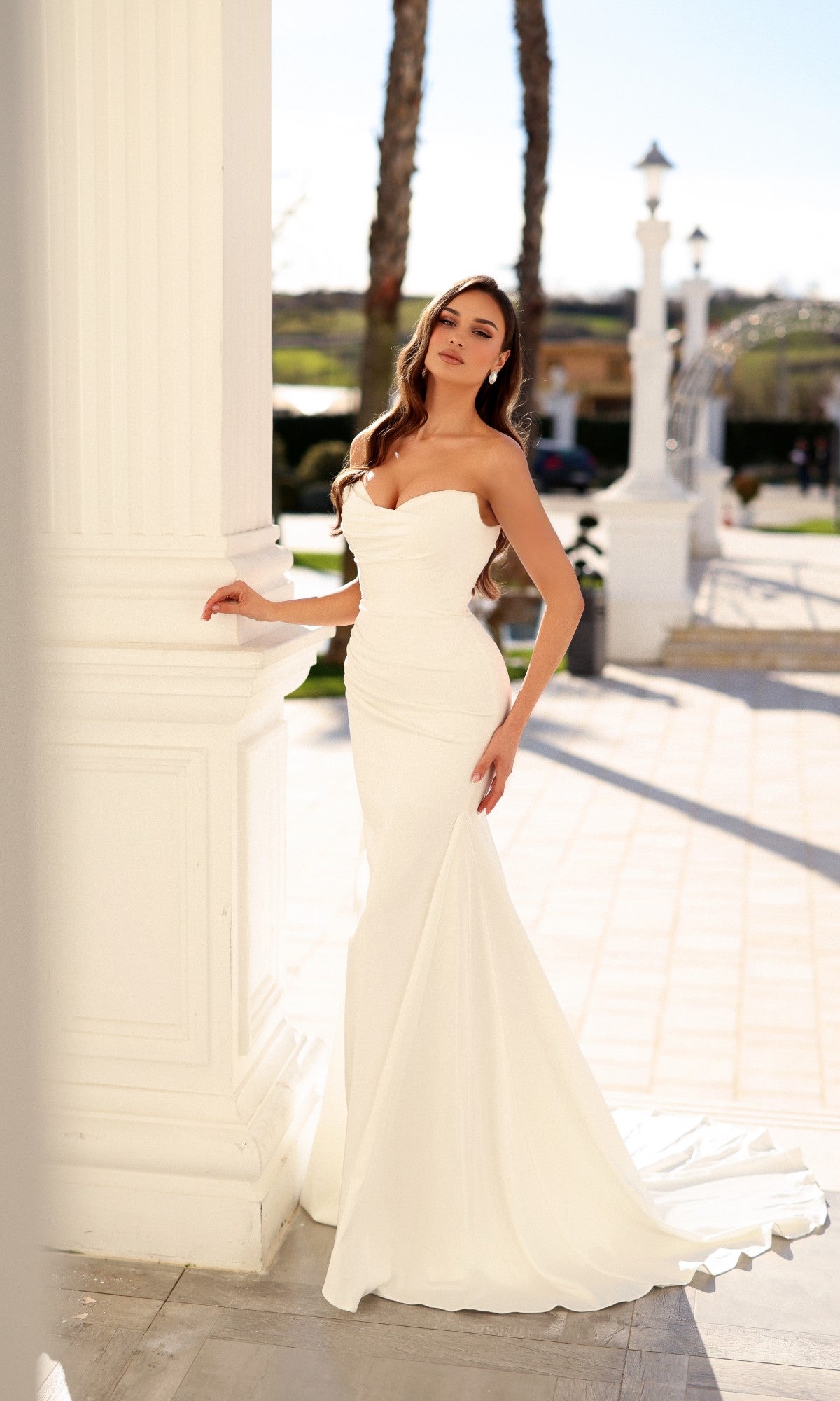 A woman wears the Tina Holly TW628 strapless, floor-length white mermaid-style bridal gown with a sweetheart neckline, leaning against a large white pillar.