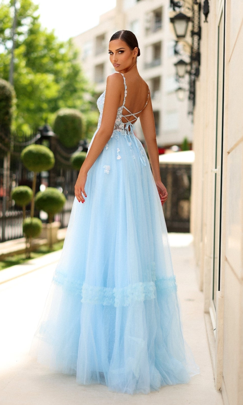 A woman in the Tina Holly TY405 long formal dress, featuring a pastel blue tulle A-line skirt and lace-up corset bodice, stands on a sunlit walkway outdoors, facing away and looking over her shoulder.