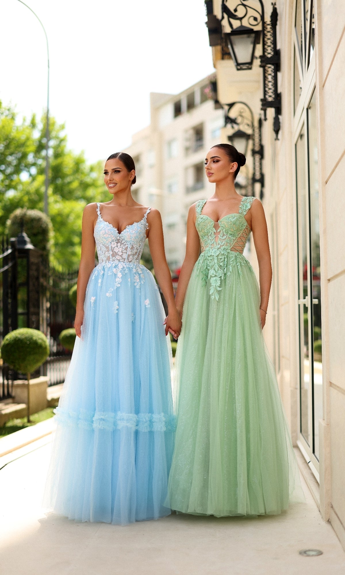 Two women stand outdoors holding hands, both wearing Tina Holly TY405 long formal dresses: one in pastel blue with a corset bodice and white floral details, the other in pale green. Both gowns have elegant floor-length tulle A-line skirts.