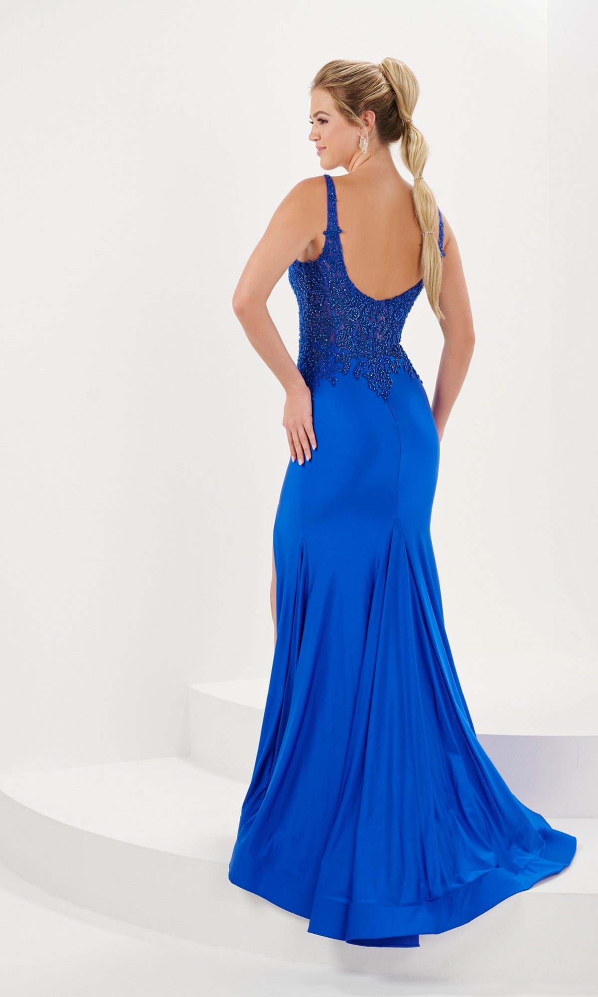 A woman poses confidently on white steps in the Long Prom Dress 16058 by Tiffany, a floor-length gown featuring a scoop neck, beaded detailing, and an open back.