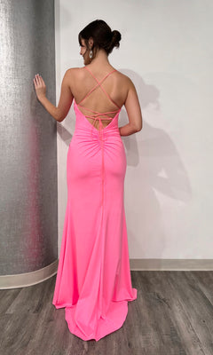 A woman in the Long Prom Dress 16069 by Tiffany, a bright pink floor-length formal jersey gown with crisscross straps, v-neckline, and open lace-up back, stands facing a wall on a wood floor.