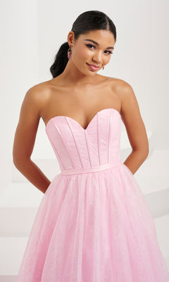 A woman poses with her hands behind her back against a plain, light-colored background, wearing the Tiffany Strapless Glitter Pastel Prom Dress 16083 featuring a shimmering corset bodice.