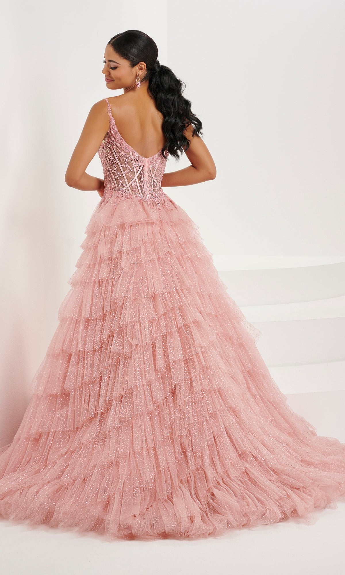 A woman poses with her back to the camera in the Tiffany Ruffled Glitter Tulle Prom Ball Gown 16099, showcasing its sparkling tulle skirt and fitted, beaded corset bodice against a plain white background.