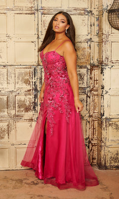 A woman poses indoors in front of a distressed paneled wall wearing the Tiffany Long Plus-Size Floral Tulle Prom Dress 16127, featuring bright pink tulle, thin straps, and a sheer overlay.
