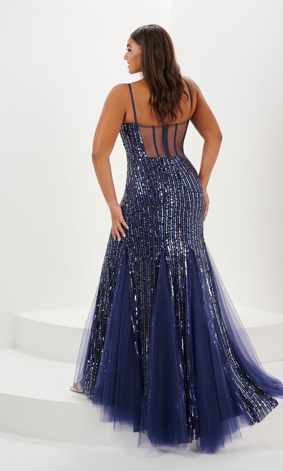 A woman in the Tiffany Plus-Size Sequin Mermaid Prom Dress 16132—navy blue with disco sequins, tulle, spaghetti straps, and a plunging v-neckline—stands on white steps, facing away from the camera.