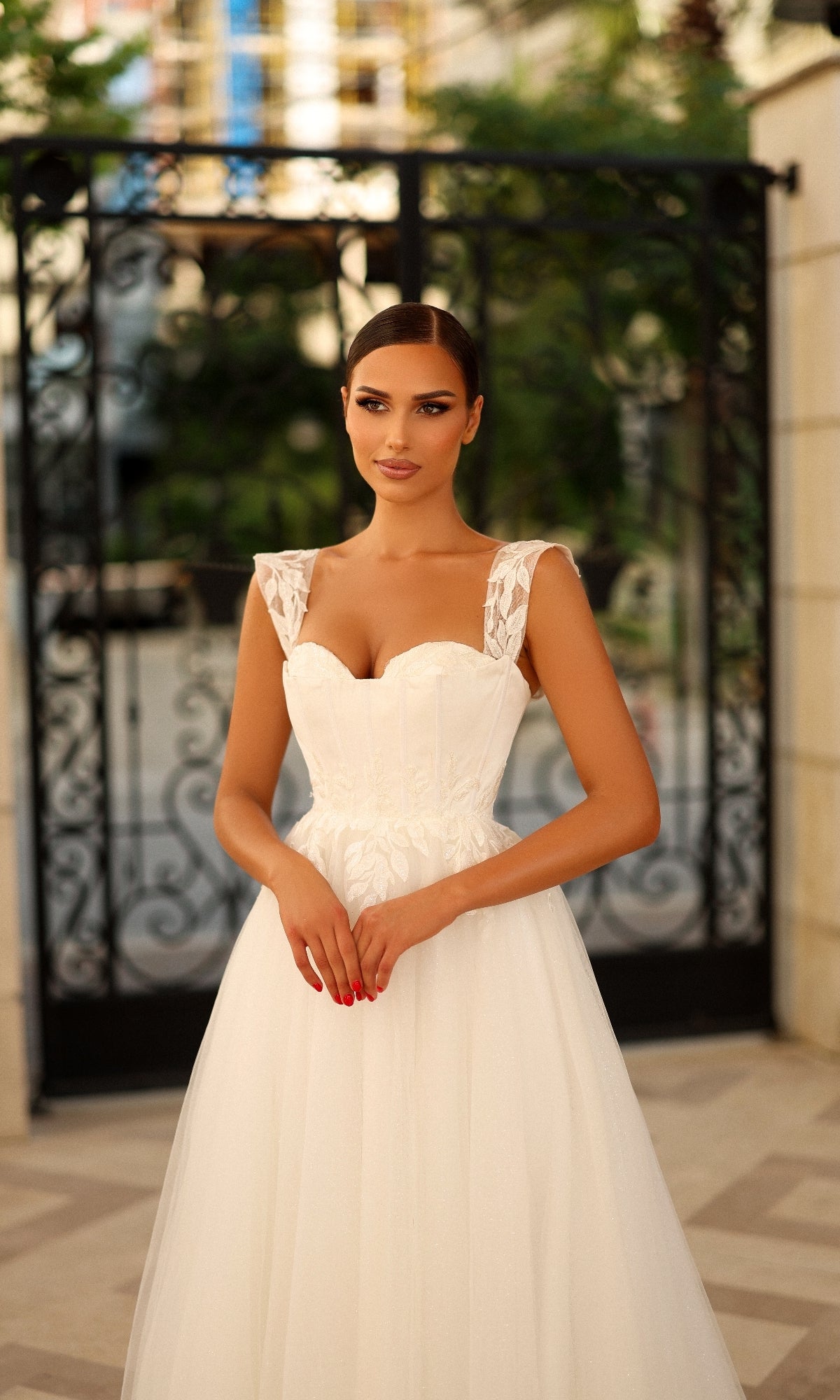 A woman wearing the Long Formal Dress: Tina Holly TW903, a white bridal gown with lace appliques and a sleeveless sweetheart neckline, stands outdoors in front of a black wrought iron gate.