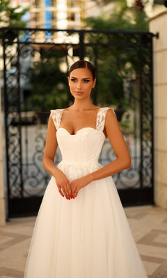 A woman wearing the Long Formal Dress: Tina Holly TW903, a white bridal gown with lace appliques and a sleeveless sweetheart neckline, stands outdoors in front of a black wrought iron gate.