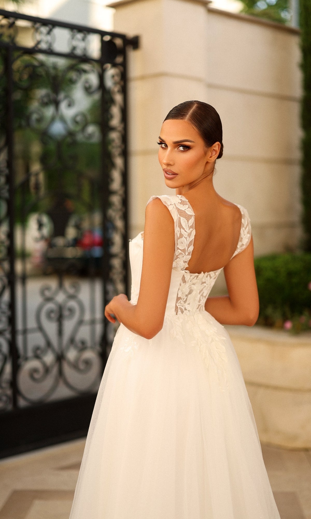 A woman in the Tina Holly TW903 long formal dress with lace appliques stands outdoors, glancing over her shoulder at the camera. Ornate black ironwork and stone walls form the background.