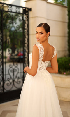 A woman in the Tina Holly TW903 long formal dress with lace appliques stands outdoors, glancing over her shoulder at the camera. Ornate black ironwork and stone walls form the background.