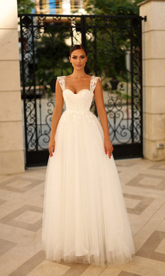 A woman stands before a black wrought iron gate wearing the Tina Holly TW903 long formal dress, featuring a fitted bodice, full tulle skirt, and delicate lace appliques.