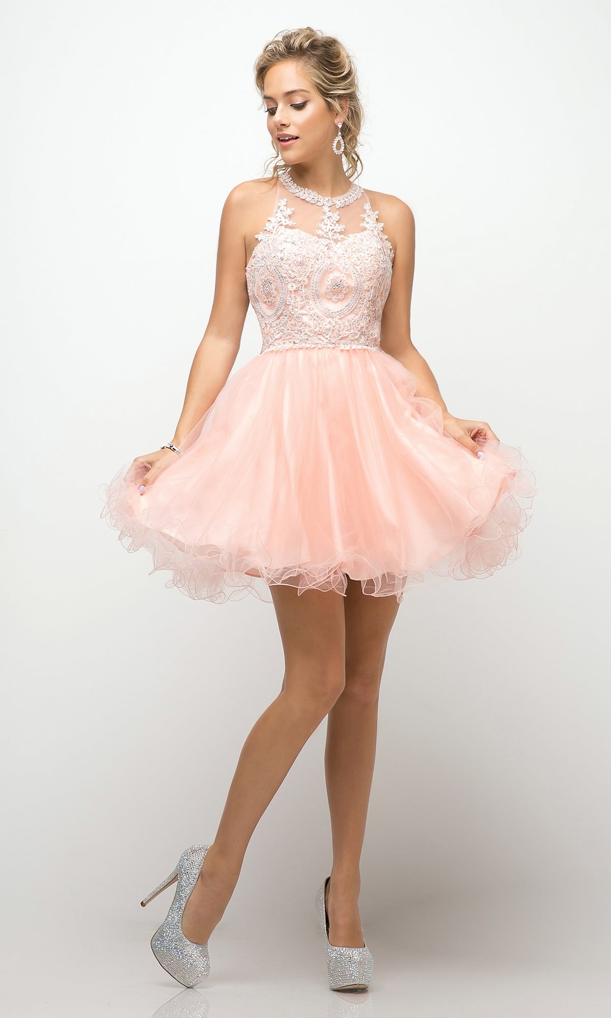 A woman in the High-Neck Short Babydoll Homecoming Dress UJ0119—a sleeveless, light pink tulle dress with an embroidered-lace bodice and silver heels—stands against a plain white background.