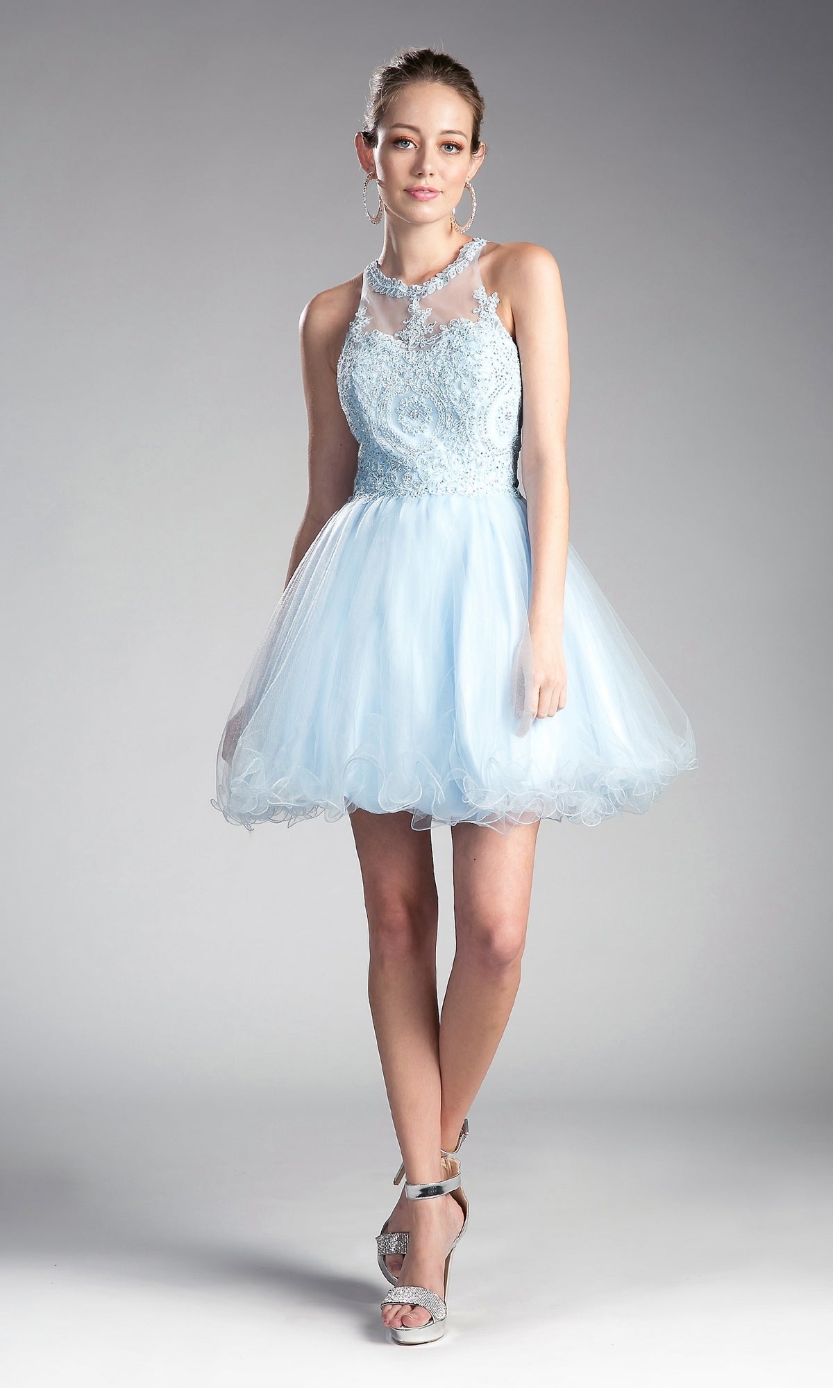 A woman in the High-Neck Short Babydoll Homecoming Dress UJ0119, a light blue sleeveless knee-length dress with an embroidered-lace bodice, pairs it with silver heels and hoop earrings against a gray background.