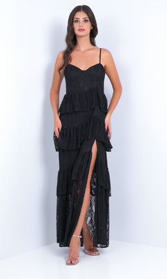 A woman models the Vanna by Velvi: a black ruffled tiered long lace prom dress with spaghetti straps and a front slit, set against a plain white background.