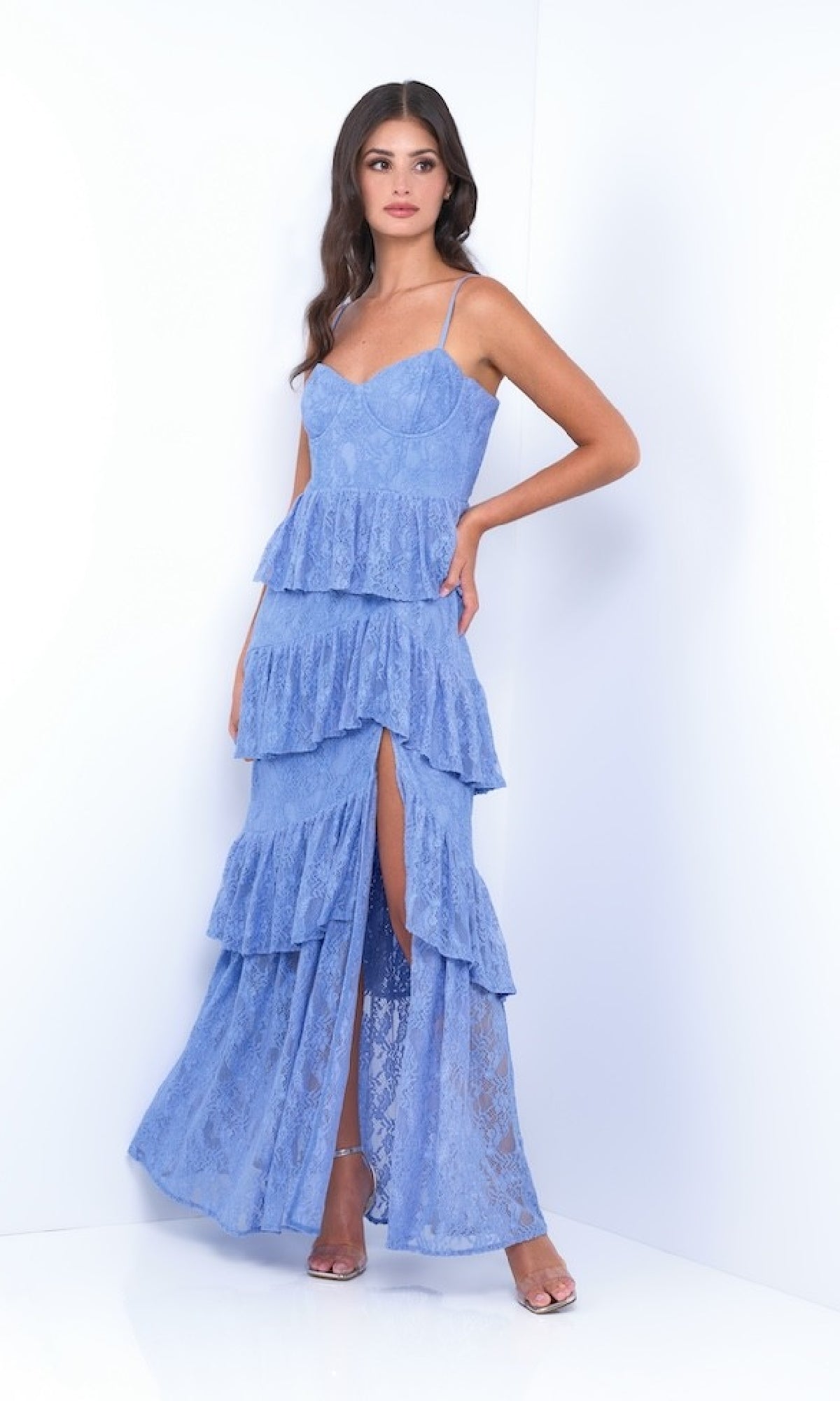 A woman models the Vanna by Velvi - a ruffled, tiered long lace prom dress with spaghetti straps and a high leg slit, shown in light blue against a plain white background.