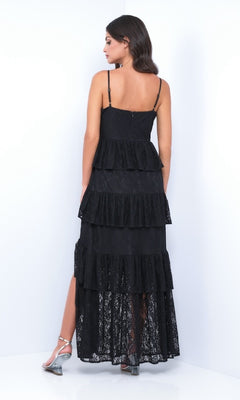A woman faces away in the Vanna by Velvi, a sleeveless, ruffled tiered long lace prom dress with a high slit, styled with silver heels against a plain white background.