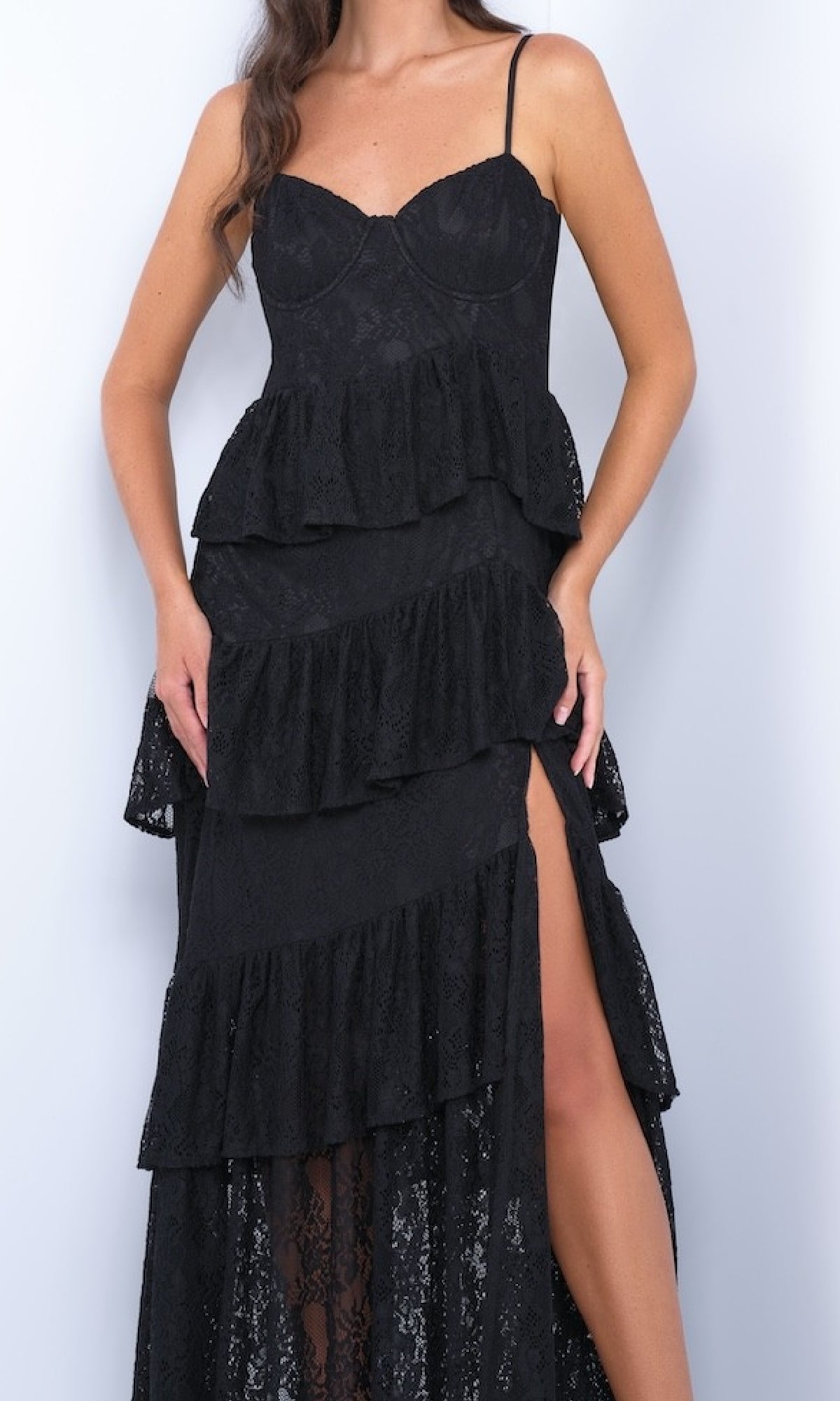 A woman in the Vanna by Velvi Ruffled Tiered Long Lace Prom Dress, sleeveless and black with a thigh-high slit, stands against a plain light background.