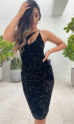 A woman poses in the Venice One-Shoulder Short Black Sequin Party Dress on a tiled floor, touching her hair, with potted plants in the background.