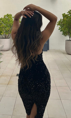 A woman with long wavy hair stands with her back to the camera and arms raised, wearing the Venice One-Shoulder Short Black Sequin Party Dress in an outdoor tiled area surrounded by potted plants.