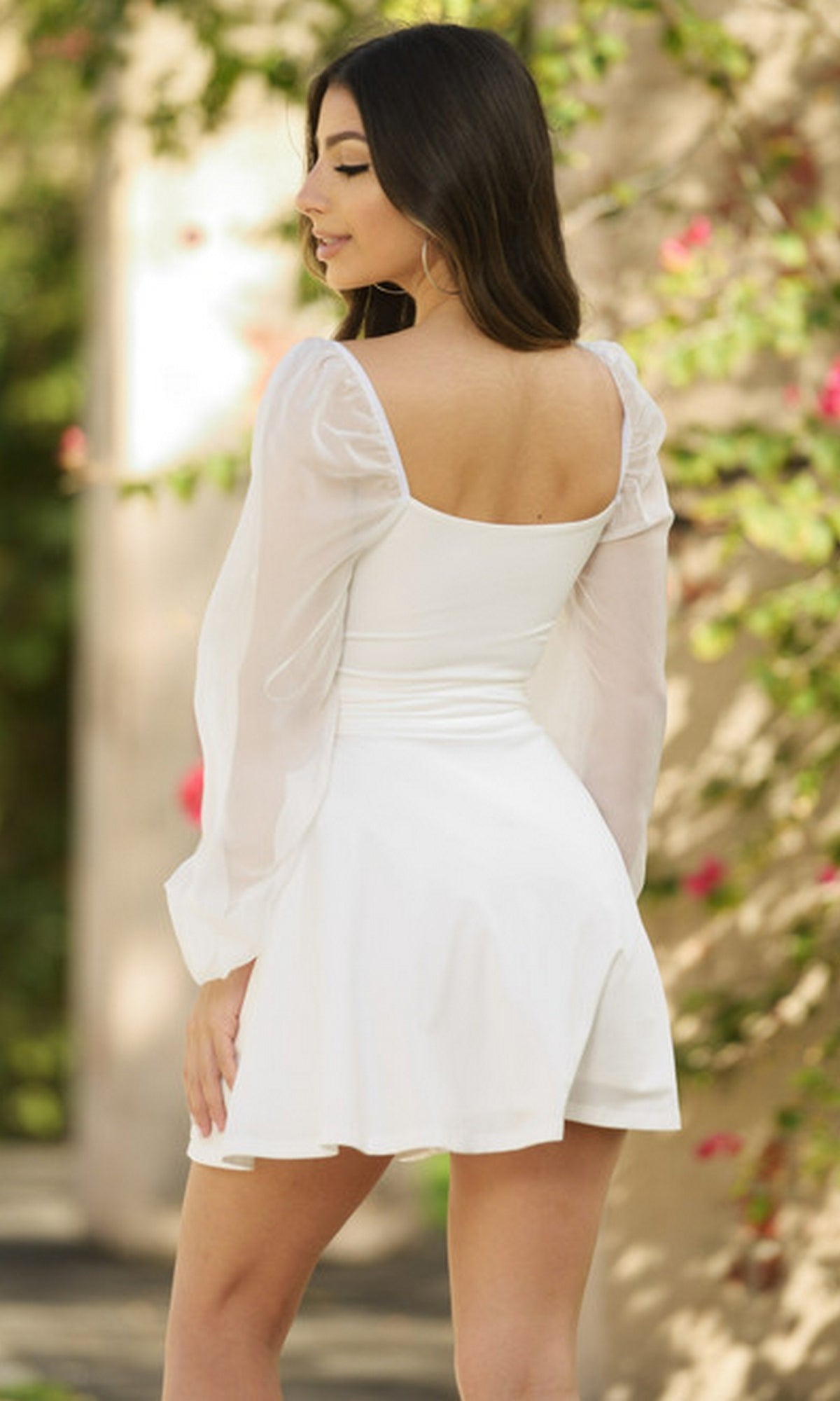A woman with long dark hair, wearing the Vera Long Sleeve Short Party Dress with sheer sleeves, stands outdoors facing away amid greenery and flowers.