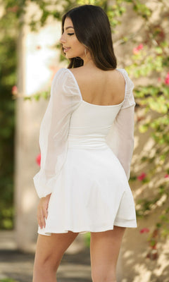 A woman with long dark hair, wearing the Vera Long Sleeve Short Party Dress with sheer sleeves, stands outdoors facing away amid greenery and flowers.
