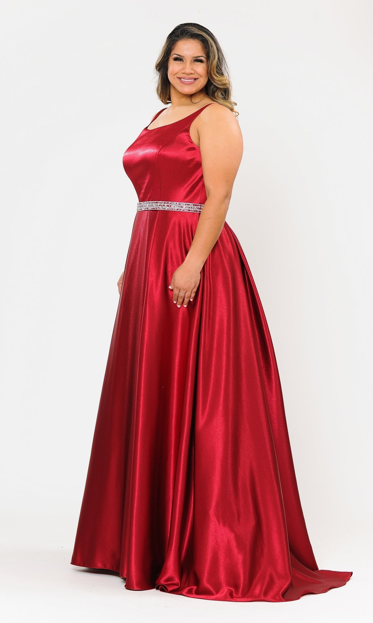 A woman wearing the Sleeveless Plus-Size Long Belted Prom Dress W1010 in shiny red with a jeweled waistband stands smiling against a plain white background.